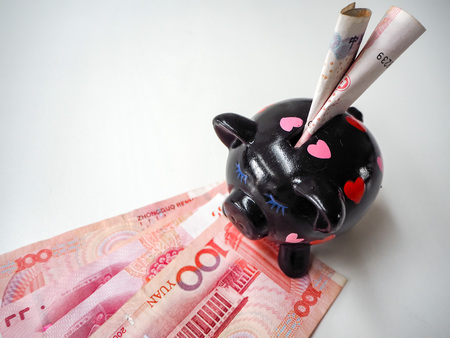 Black piggy bank standing next to Chinese 100 rmb banknotes on a white wooden background. The pig symbols fortune in the Chinese zodiac.の写真素材