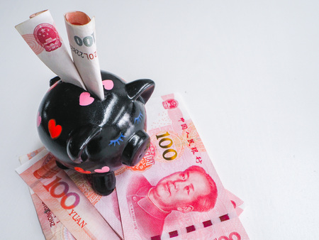Black piggy bank standing next to Chinese 100 rmb banknotes on a white wooden background. The pig symbols fortune in the Chinese zodiac.の写真素材