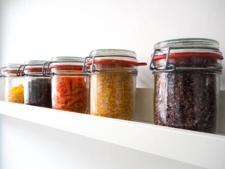 Collection of rustic glass jars filled with dry food such as pasta, lentils and risotto on a white shelf following a zero waste lifestyleの写真素材