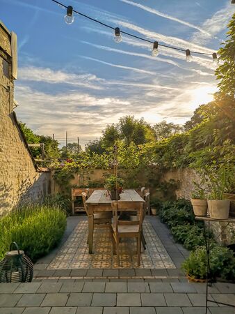 Vintage cement tiles in a small walled urban garden full with green plants and wooden furniture in...