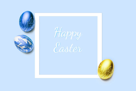Frame and three easter eggs decorated with gold and blue color on blue background. Happy Easter title. Flat lay, top view, copy space.の写真素材