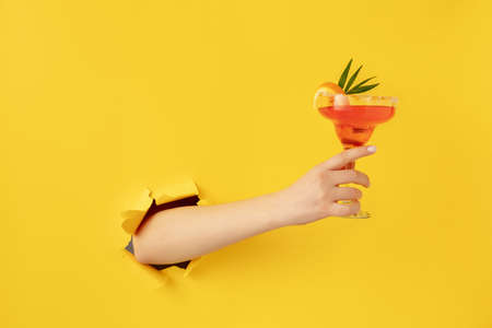 Female hand with cocktail glass breaks through yellow paper background.の写真素材