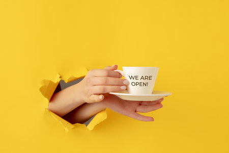 Female hand with cup of tea or coffee torn through yellow paper background.