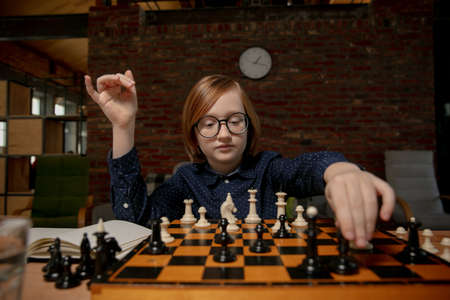 Little boy in eyeglasses sitting at home and playing chess alone.の写真素材