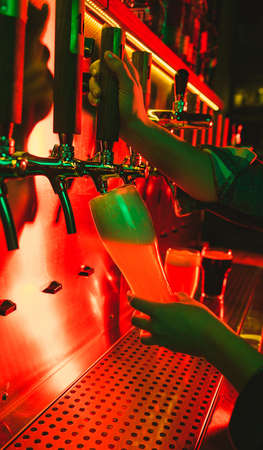 Young woman barman pouring a cold lager beer from tap to glass in neon light.の写真素材