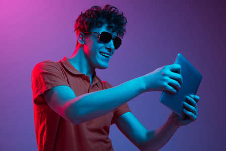 Young man in sunglasses with device isolated on gradient background in neon