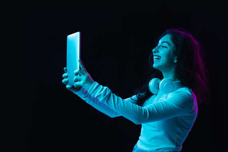 Young beautiful girl using device isolated on dark background in neon lightの写真素材