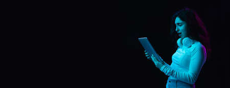 Young beautiful girl using device isolated on dark background in neon lightの写真素材