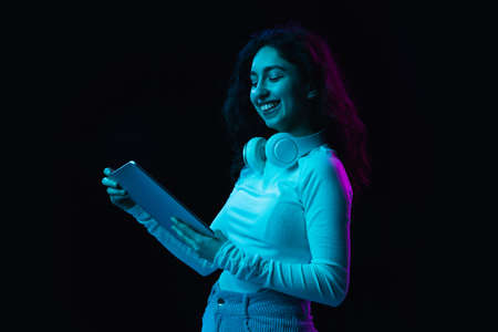 Young beautiful girl using device isolated on dark background in neon lightの写真素材