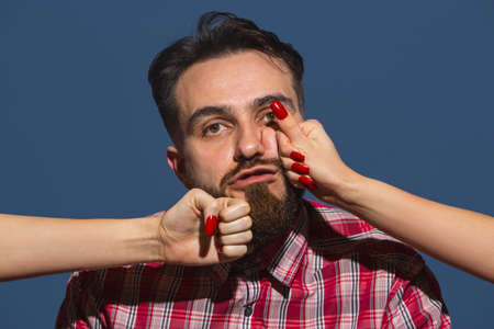 Portrait of young man with female hands punchning his face on blue backgroundの写真素材