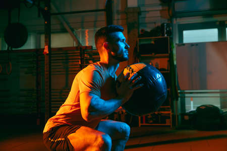 Young athletic man, fitness instructor training in gym in mixed neon light.