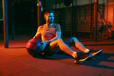 Young athletic man, fitness instructor training in gym in mixed neon light.