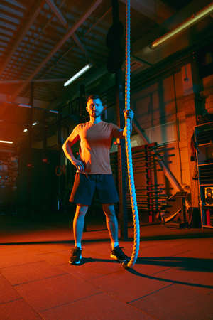 Young athletic man, fitness instructor training in gym in mixed neon light.
