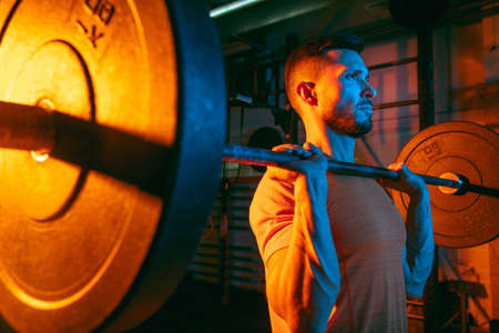 Close up young athletic man training with barbell in gym in mixed neon light.の写真素材