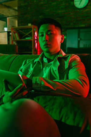 Young Korean man using tablet at home interior in gradient red green neon lightの写真素材