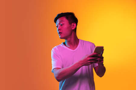 Portrait of Asian young man with smartphone isolated over orange yellow background in neon light.の写真素材