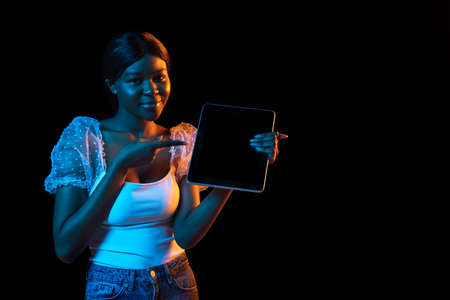 Young African beautiful girl with tablet isolated over dark background in mixed neon light.の写真素材