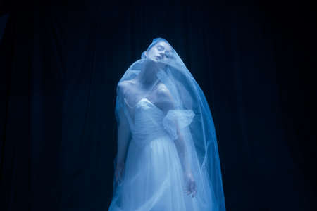 Despair. Young graceful balerina in image of ghost bride in art performance isolated on dark background in neon light.の写真素材
