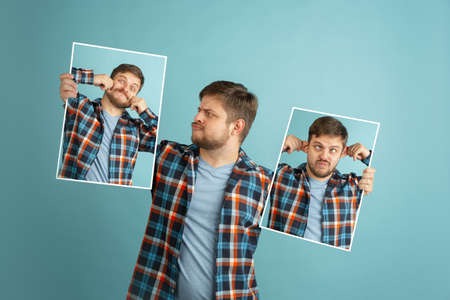 One young man in casual clothes holding his portraits with different emotions isolated on light blue...