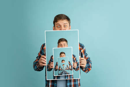 Mirror effect. Young man in casual clothes holding his portrait isolated on light blue background....