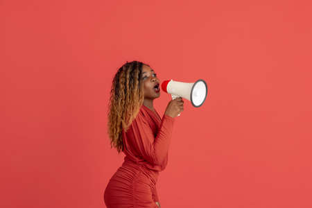 Young attractive african girl with loudspeaker in red fashionable dress isolated on coral red background. Human emotion conceptの写真素材