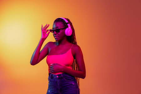 Young pretty African girl, female fashion model in sunglasses and white headphones isolated on yellow background in neon light.の写真素材