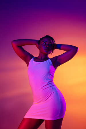Beautiful African girl in white short summer dress posing isolated over bright colorful neon...