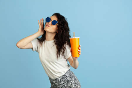 Portrait of pretty young curly caucasian girl in sunglasses with paper cup isolated over light blue...