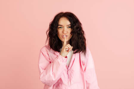 Studio image of young smiling Caucasian woman with finger to lips looking at camera posing on light...