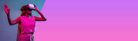 Portrait of young african woman in vr glasses on bright pink and grey background. Concept of virtual...