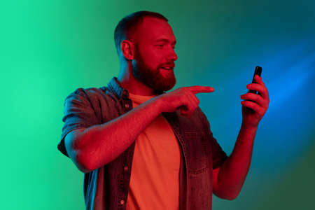 Studio image of young caucasian bearded man with smartphone isolated on gradient green blue...