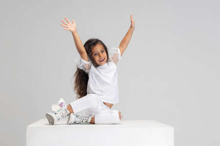 Portrait of little cute smiling Caucasian girl sitting on big box with hands up isolated over white...
