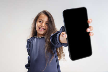 Showing device screen. One smiling preschool girl in blue dress holding smartphone isolated over white studio background.の写真素材
