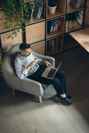Vertical image of serious young businessman sitting in comfortable armchair with laptop and cup of coffee. View from aboveの写真素材