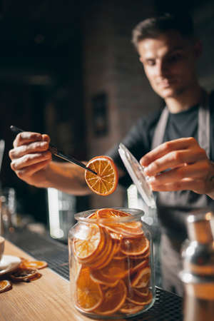 One young handsome barista in apron serves hot aromatic coffee pulling slices of dried orange from jarの写真素材