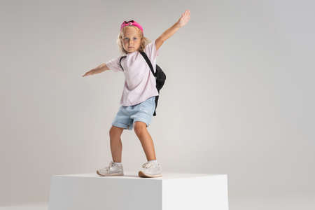 One little preschool Caucasian girl in casual clothes with backpack stands on big box isolated over white studio background.の写真素材