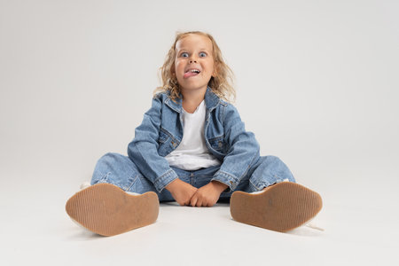 Portrait of little smiling preschool Caucasian girl in denim clothes sitting on floor isolated over white studio background.の写真素材