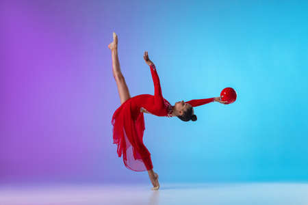 Young girl, rhythmic gymnast in red dress with ball in motion and action isolated on purple blue gradient neon background.の写真素材