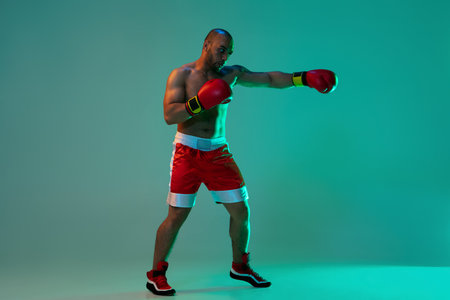 Side view. One professional boxer in red gloves training, exercising over blue green background in neon light.の写真素材