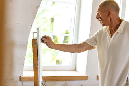 Mature or senior grey painter with a brush standing in front of camera. Serious man in a paint-stained shirt working at studio or homeの写真素材