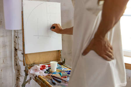 Mature or senior grey painter with a brush standing in front of camera. man in a paint-stained shirt working at studio or homeの写真素材