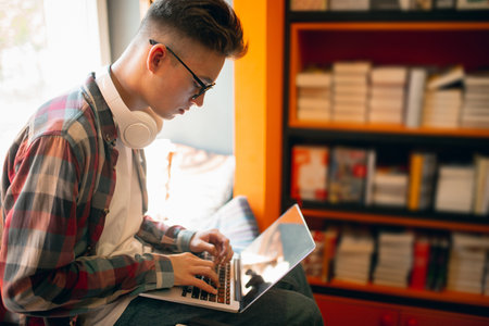 Studying, doing homework. One young caucasian man, student in glasses sitting with laptop at home or library background. Education, studying and student life concept.の写真素材