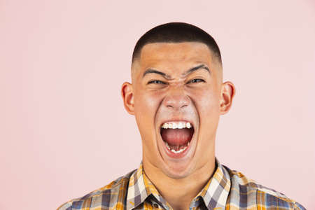 Close-up portrait of young screaming, shouting asian man, guy in a plaid shirt isolated over light pink studio background. Concept of human emotions, leisure, youth.の写真素材