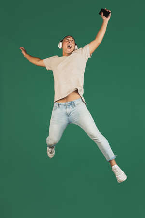 Portrait of young handsome asian man in headphones jumping with phone isolated over dark green...