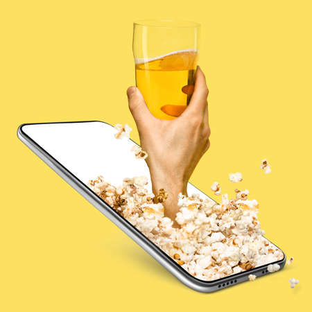 Creative collage. Male hand holding glass of beer sticking out of smartphone screen over yellow background.の写真素材