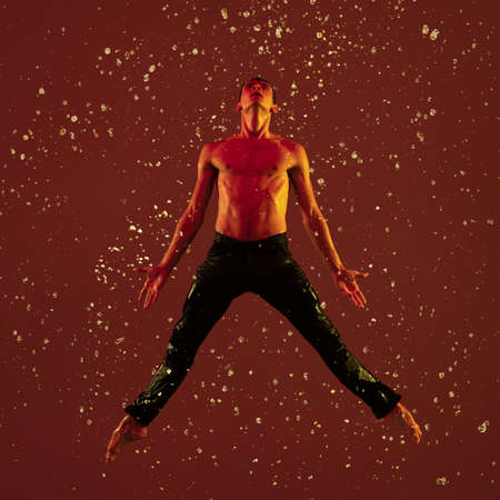 One young male ballet dancer in motion, action under the rain over red background in neon light.