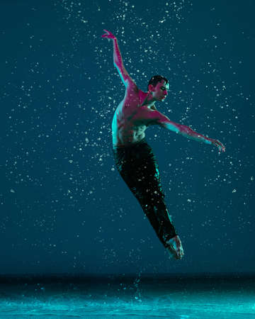 One young male ballet dancer in motion, action under the rain over blue background in neon light.の写真素材