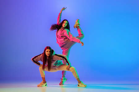 Two young beautiful women in casual clothes dancing brakedance, hip-hop over blue background in neon light.の写真素材