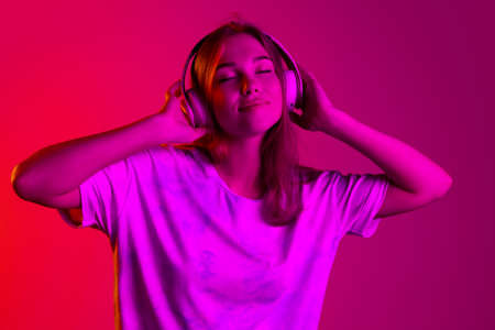 Beautiful young caucasian girl with with closed eyes listening to music in headphones isolated on pink background in neonの写真素材