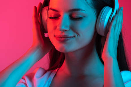 Close-up cropped portrait of young beautiful girl with headphones on gradient pink red studio backgroundの写真素材
